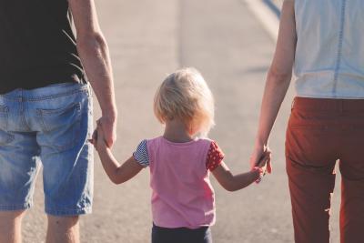 Photo taken from behind of a child holding the hands of their parents. 