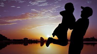 Photo of a sunset showing the silhouette of a father lifting his young son up in the air.