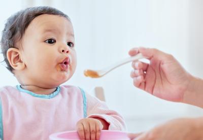 Baby eating solid baby food from a spoon being held by caretaker.