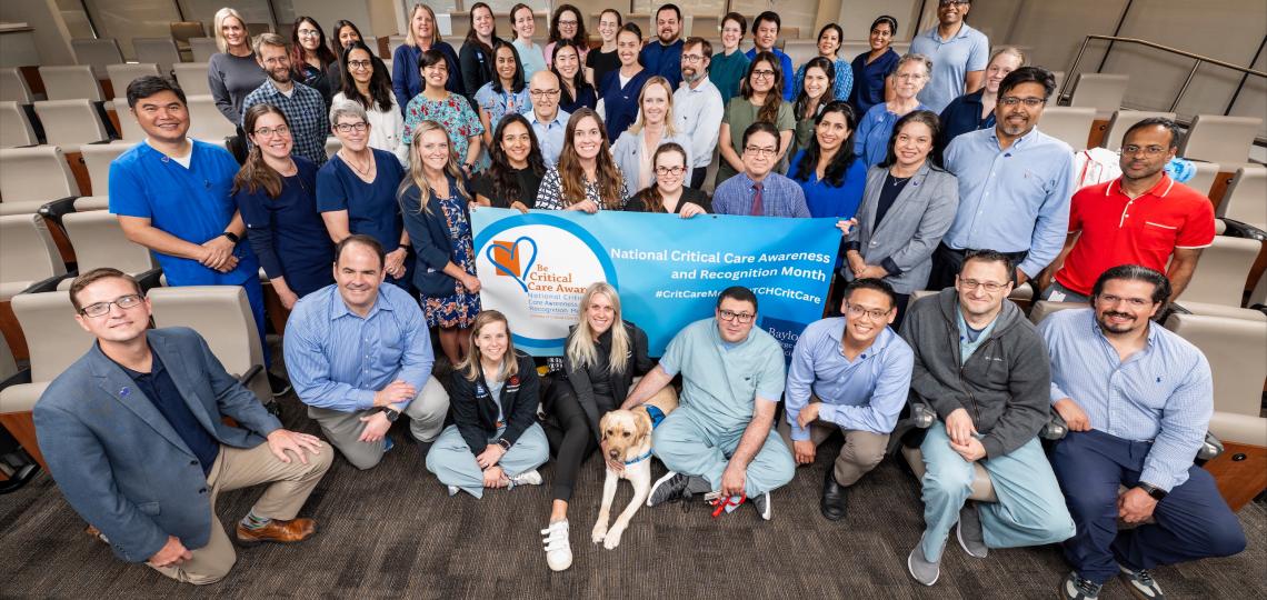 A large group of faculty members smiling while holding a banner reading National Critical Care Awareness and Recognition Month. There is also a golden lab dog in the front row.