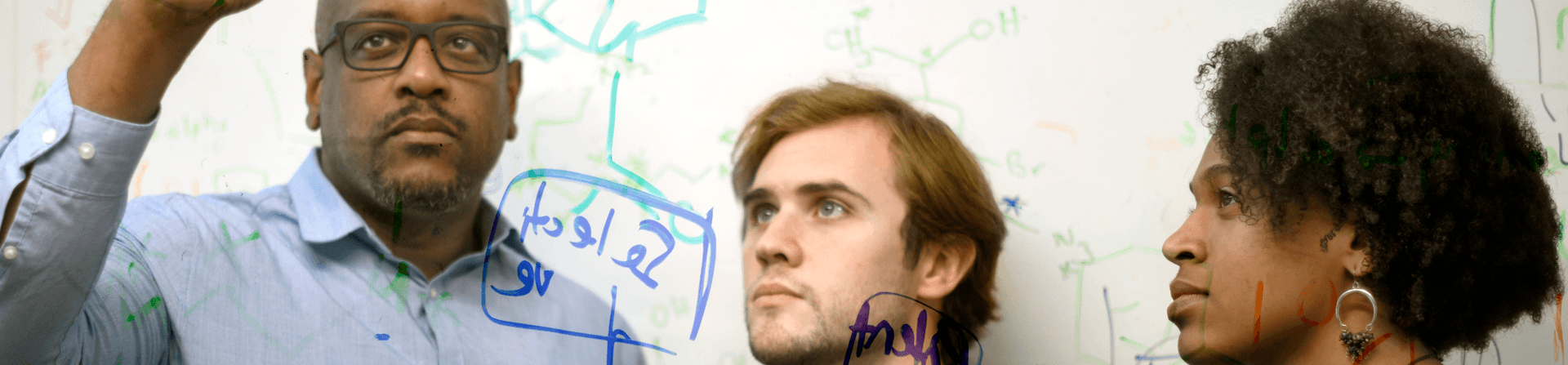 Three scientists write equations on a clear board