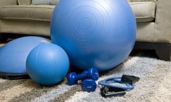 Blue exercise ball, weights and band sitting on the floor in front of a grey couch. 