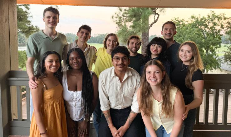 Eleven people smiling on a porch, with a wooded area at sunset in the background.