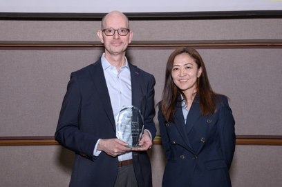 two people standing together smiling, with one holding an award plaque