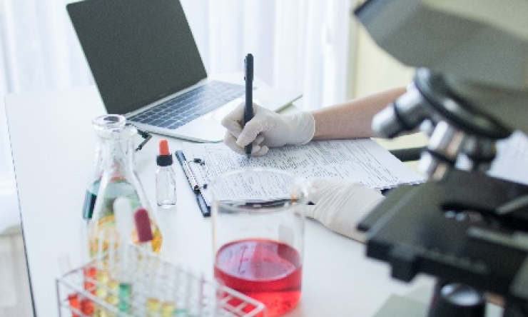 A research working in a lab with microscope and a lab top completing paperwork.