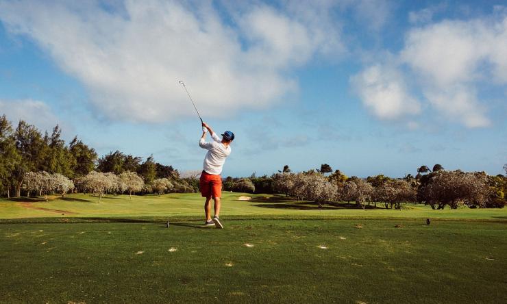 Photo of a man golfing, taken from behind as he swings with the blue sky in the background.