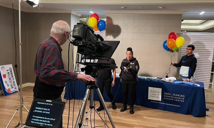 A video team behind a camera recording a person standing in front of a table with festive balloons. 