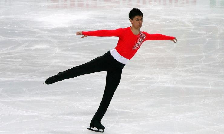 A man in black pants and a red top figure skating with arms outstretched and one leg in the air.
